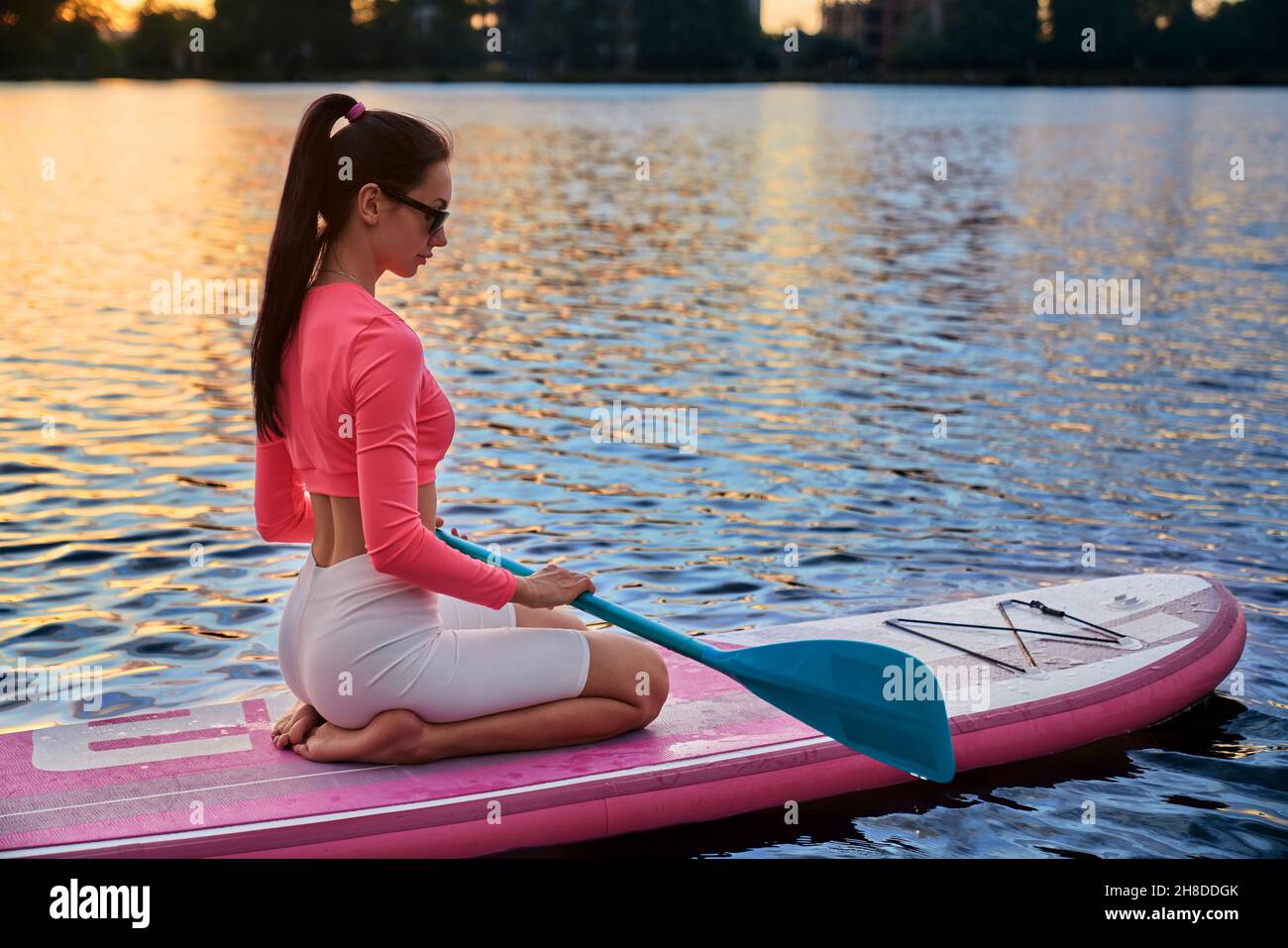 Junge Frau in Sportkleidung und Sonnenbrille sitzt auf dem Brett mit langem Paddel in den Händen. Toller Sommeruntergang auf dem lokalen See. Wassersport und Menschen Konzept. Stockfoto