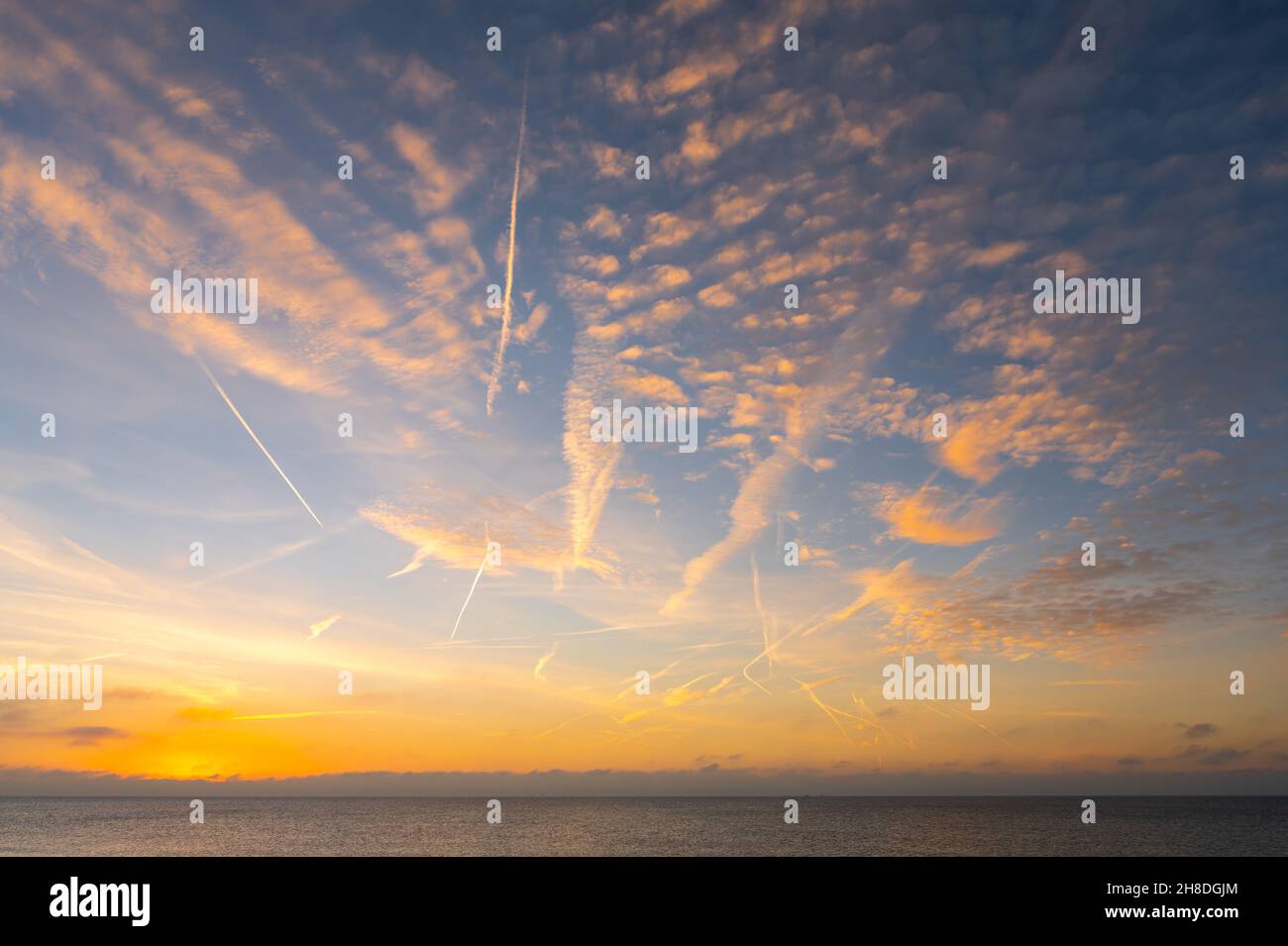 Kondensstreifen & leichte, flauschige Wolken über dem Meer, mit einer orangefarbenen Tönung, die vom morgendlichen Sonnenaufgang im Winter in Großbritannien erhellt wird. Wolken von der Sonne beleuchtet. Stockfoto