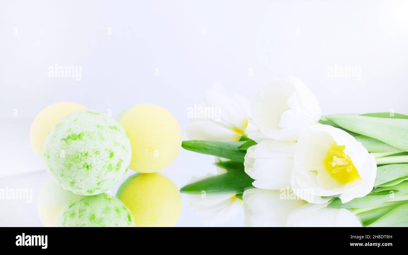 Neben weißen Tulpen wird eine grüne und gelbe Salzbombe auf einem weißgrauen Hintergrund reflektiert. Mineralisches Make-up. Banner. Wellnessbehandlung. Banner. Speicherplatz kopieren Stockfoto