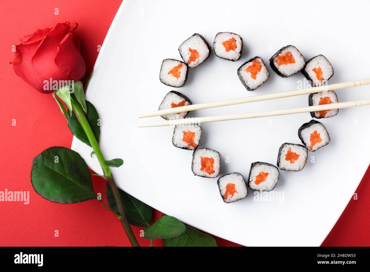 Sushi-Rollen, die in Herzform auf einem weißen Teller ausgelegt sind. Rote Rose auf rotem Hintergrund. Kreative Grüße. Das Konzept der japanischen Küche für V Stockfoto