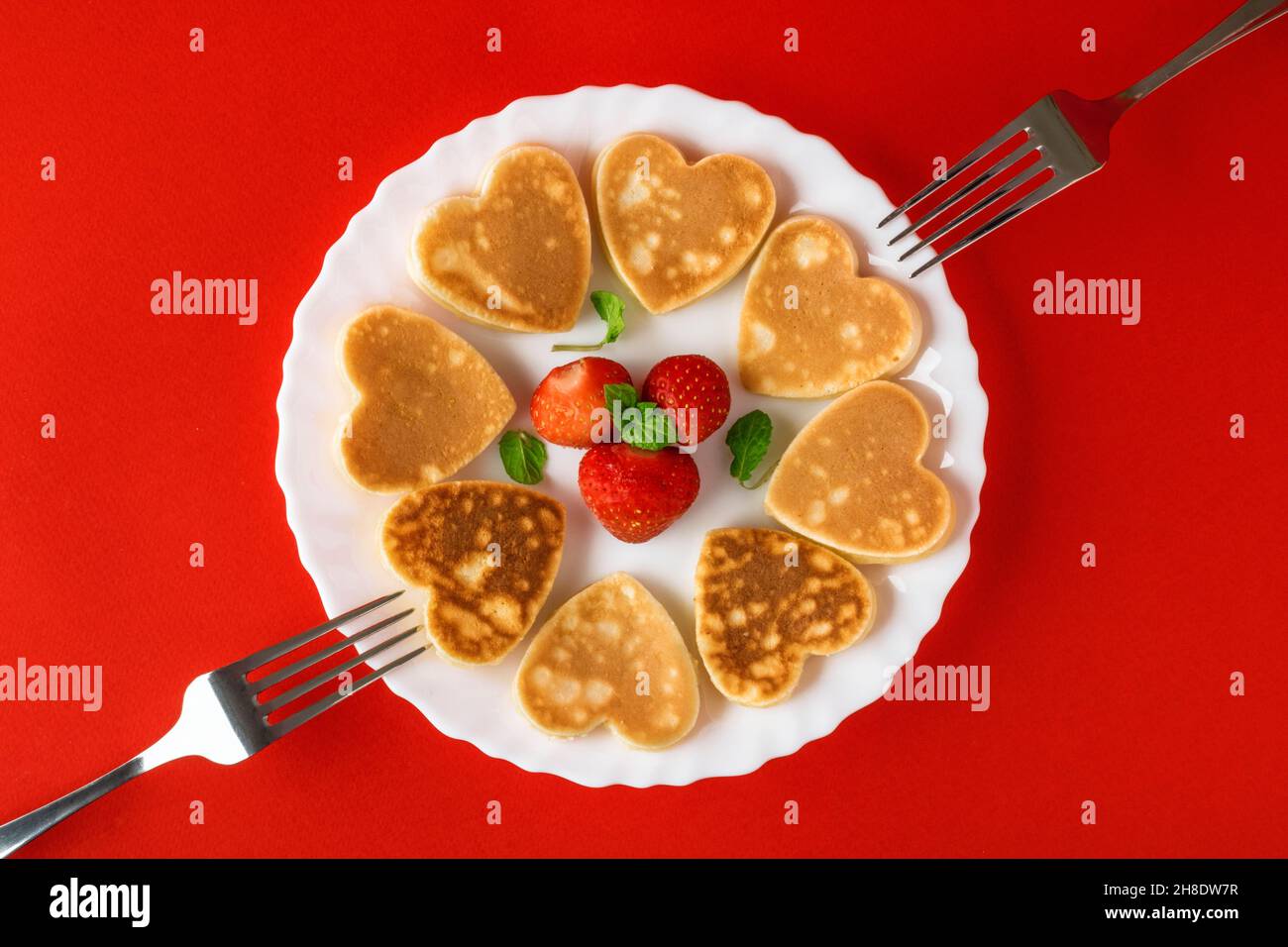 Kreatives Frühstück zum Valentinstag. Herzförmige Pfannkuchen mit Erdbeeren auf einem Teller mit Gabeln, auf rotem Hintergrund. Valentinstag Hintergrund Stockfoto