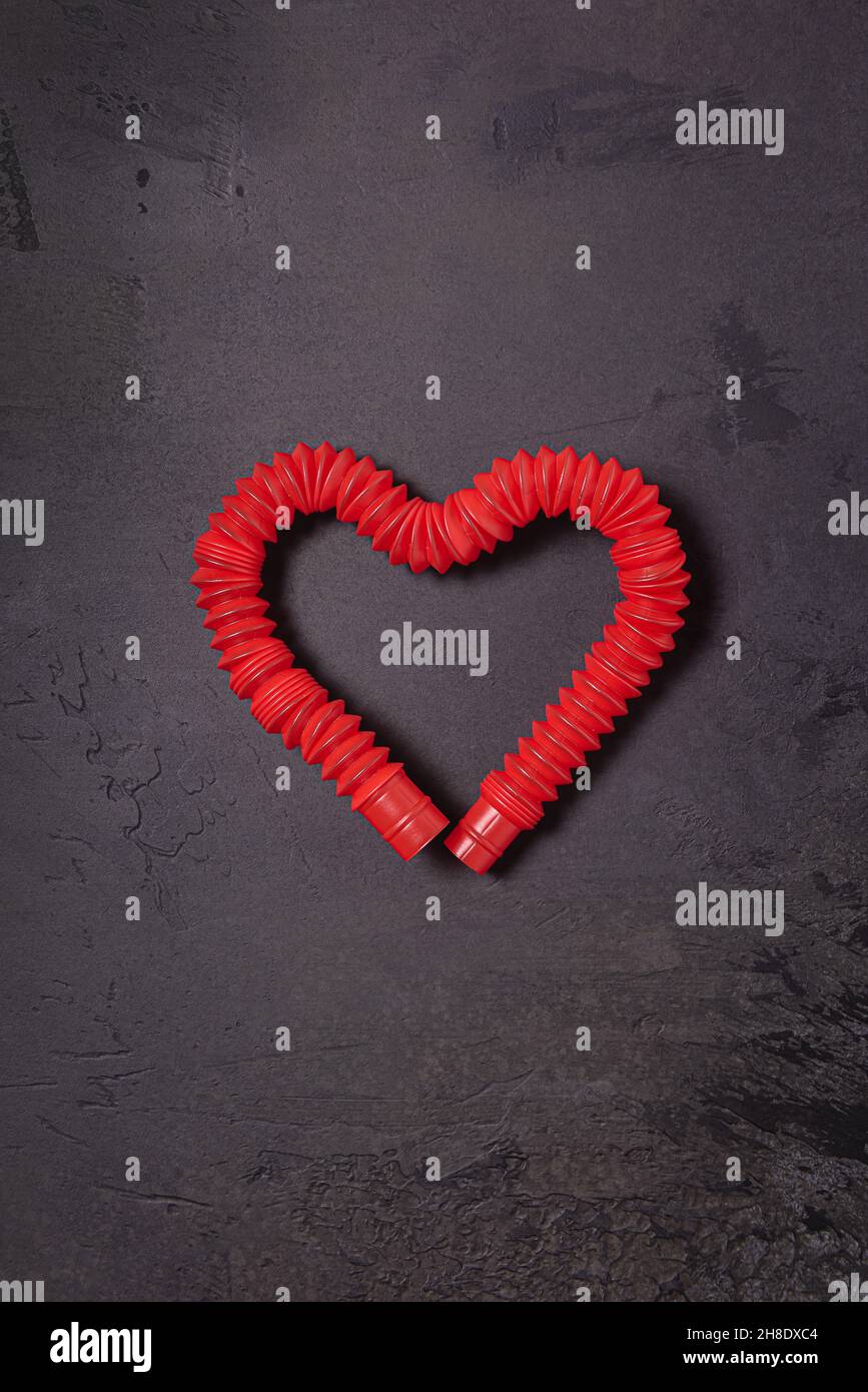Gestreckt leuchtend rot Pop it tube Kinder Spielzeug gebogen in Form von Herz. Kunststoff-Anti-Stress-Spielzeug in der Mitte der schwarzen strukturierten Stein Hintergrund. Flach liegend. C Stockfoto
