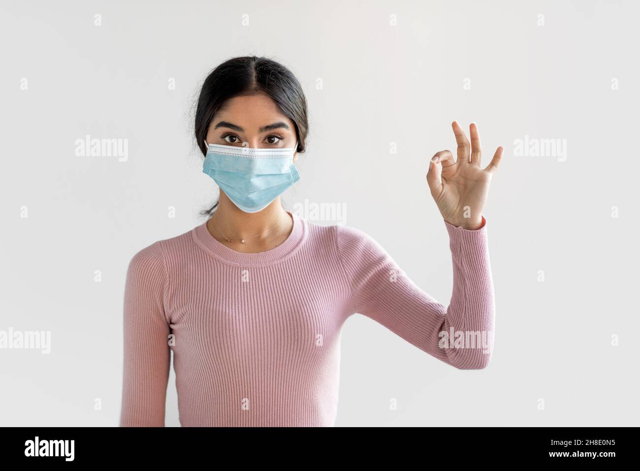Fröhliche lustige tausendjährige indische Frau in Schutzmaske zeigt ok Zeichen auf grauem Wandhintergrund Stockfoto