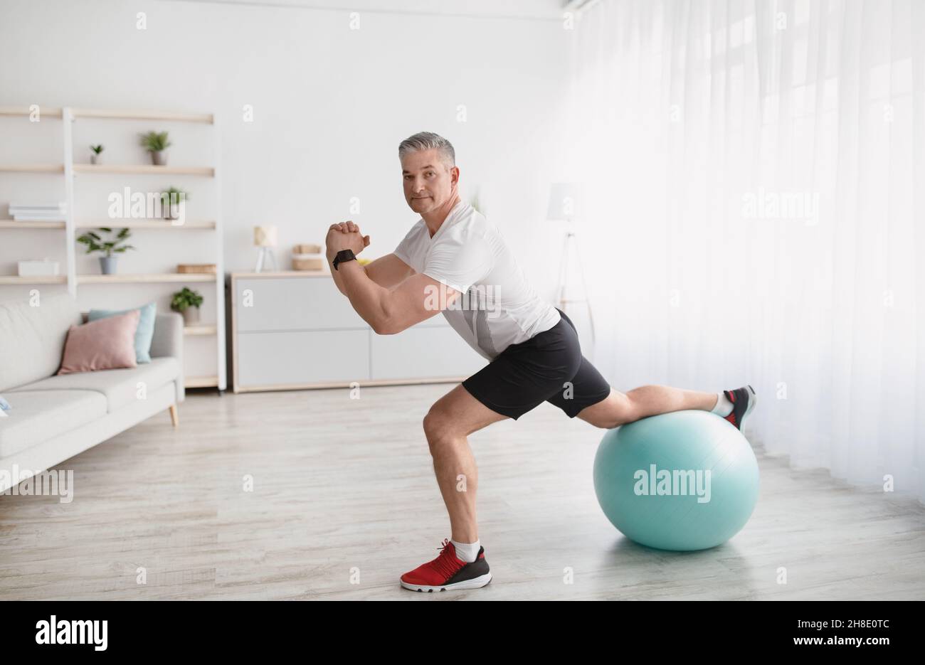 Heimtraining. Älterer Mann macht Ausfallschritte mit Fitball, trainiert zu Hause und lächelt an der Kamera, kopiert den Raum Stockfoto