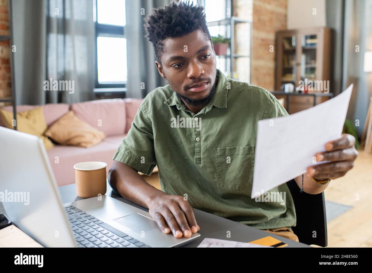 Serious junger afroamerikanischer Mann sitzt am Tisch mit Laptop im Wohnzimmer und untersucht persönliche Finanzen Stockfoto