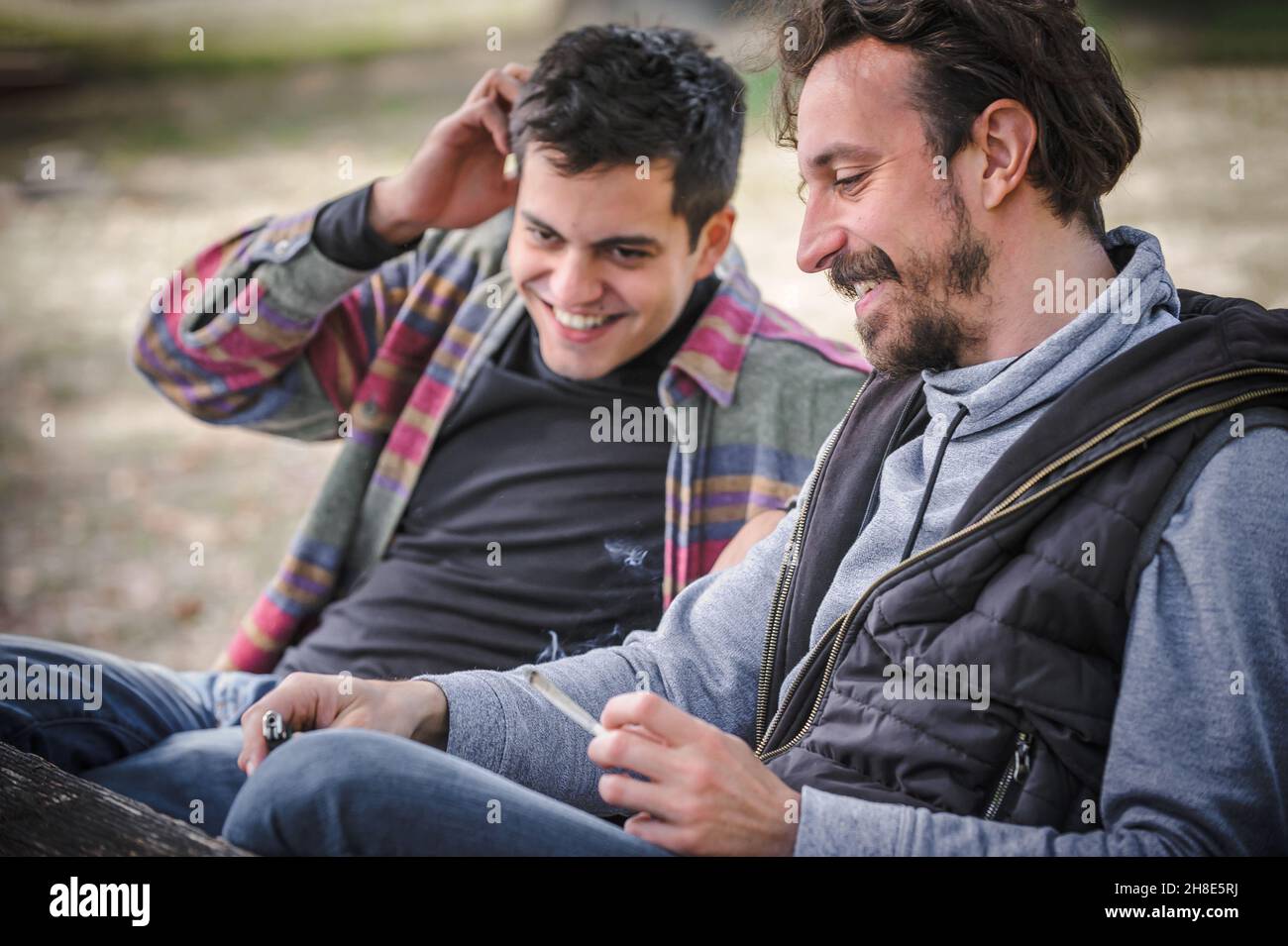 Zwei entspannte und lächelnde, glückliche junge Freunde, die Cannabis-Marihuana-Ganja oder Haschisch-Joint im öffentlichen Park im Freien rauchen. Zwei Männer rauchen thc-grünes Gras t Stockfoto