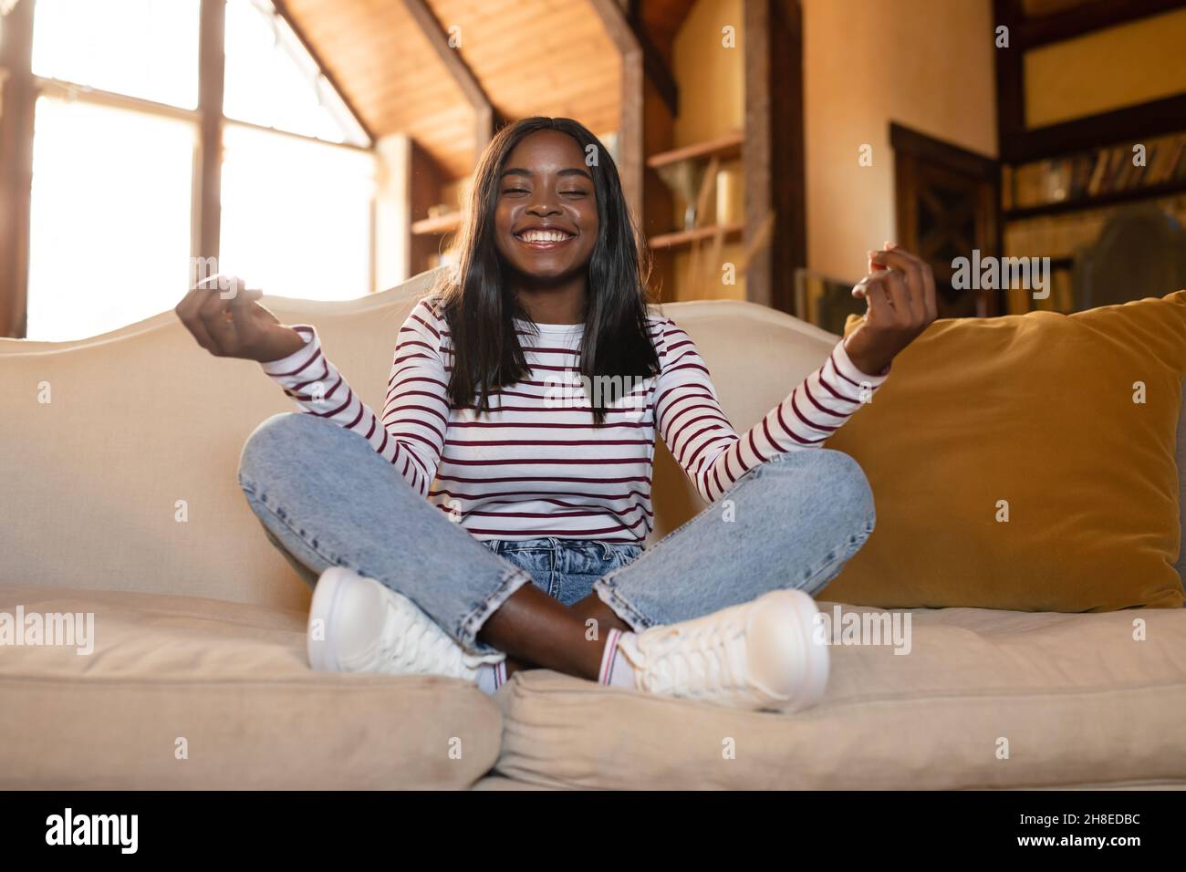 Friedliche afroamerikanische Frau, die in Lotushaltung auf der Couch sitzt, Zen-Yoga praktiziert und zu Hause meditiert Stockfoto