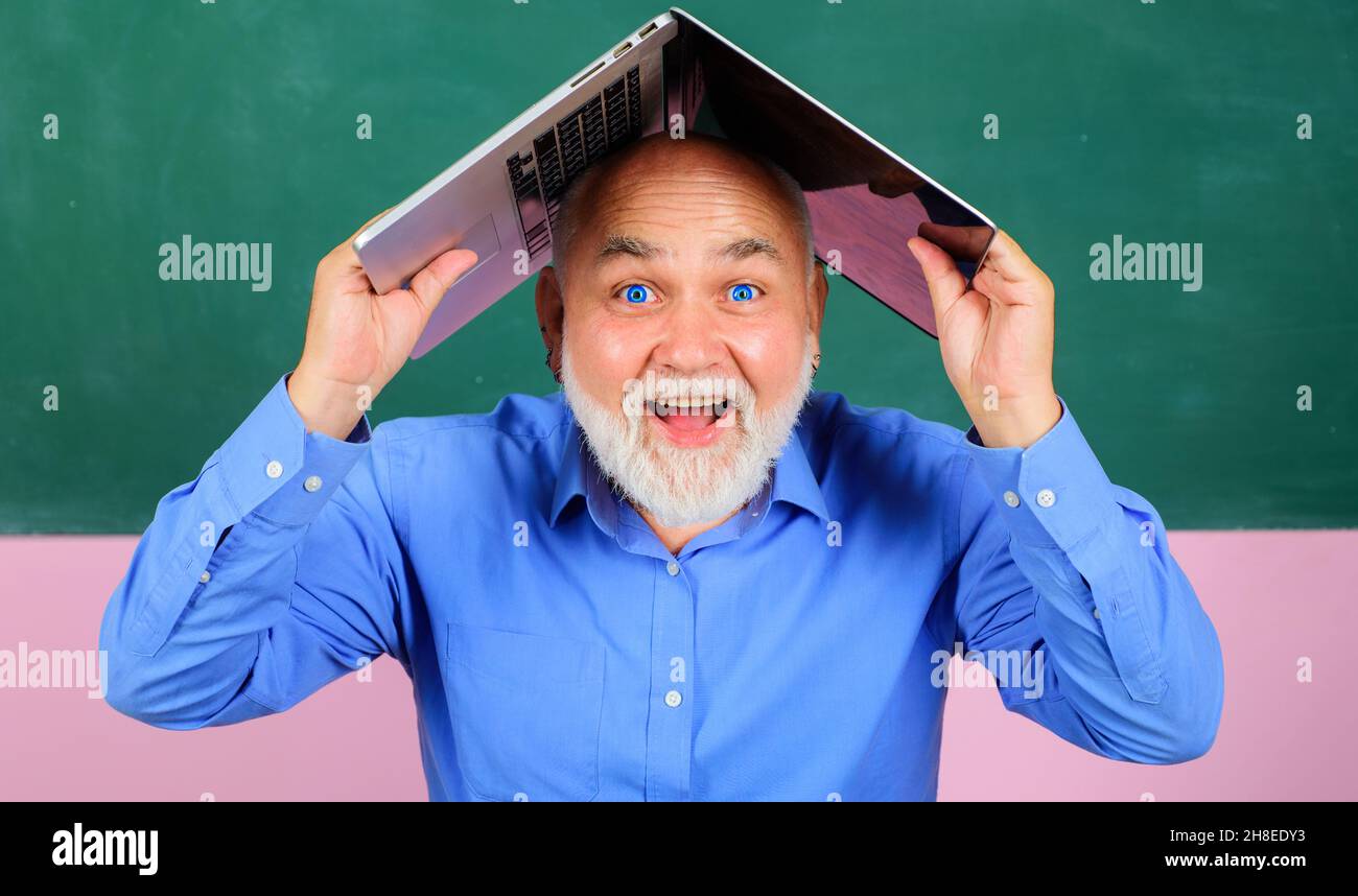 Glücklicher bärtiger Mann mit Laptop über dem Kopf. Lächelnder Lehrer mit Notizbuch im Klassenzimmer Stockfoto
