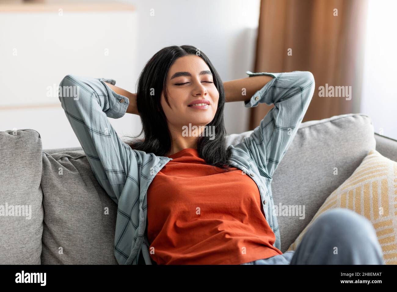 Entspannte junge arabische Frau, die sich auf der Couch zurücklehnte, sich mit den Händen hinter dem Kopf auf dem bequemen Sofa ausruhte und ihre Augen schloss Stockfoto