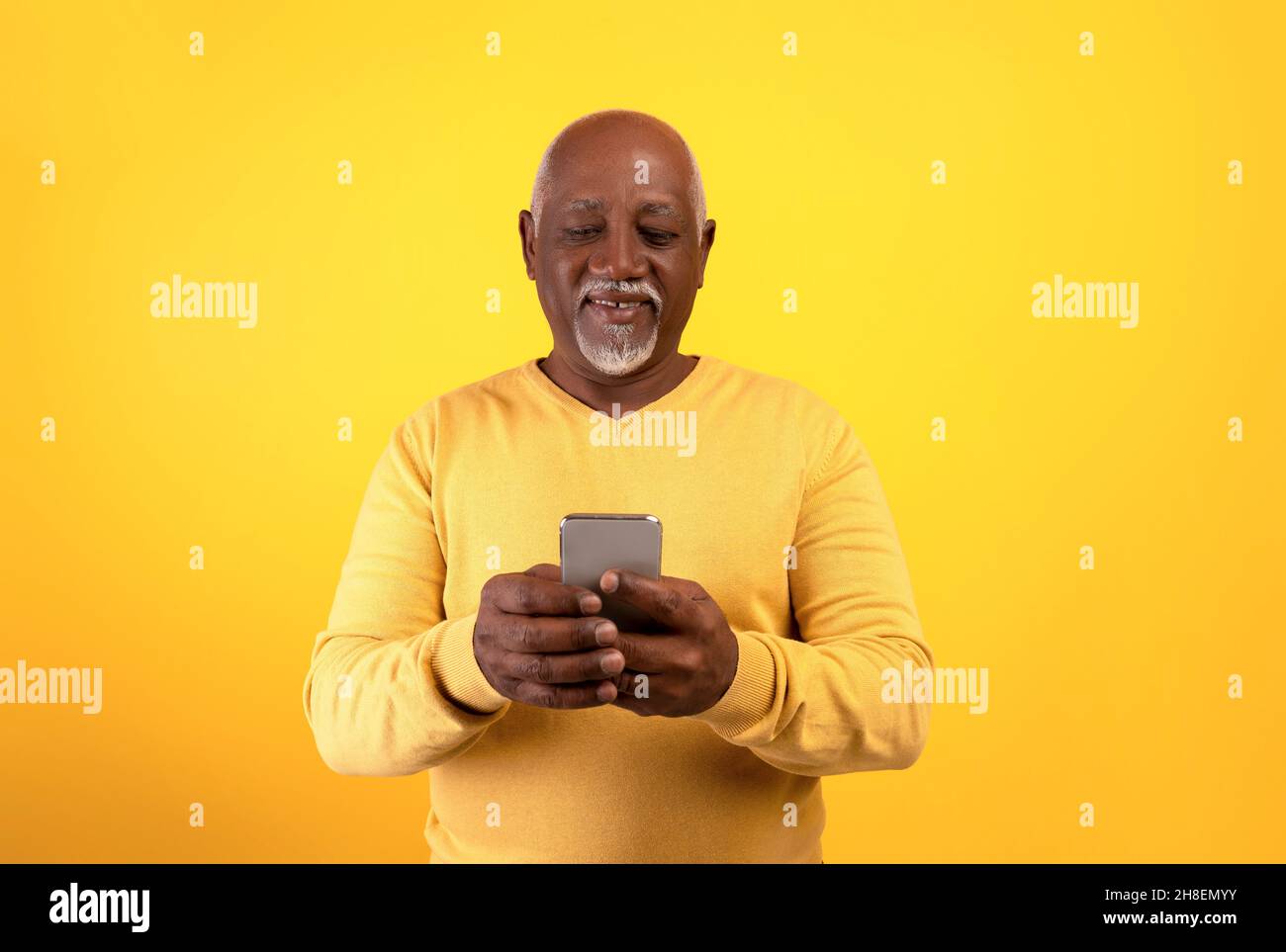 Glücklicher älterer schwarzer Mann mit Smartphone, SMS an die Familie, SMS senden, im Web auf orangefarbenem Studiohintergrund surfen Stockfoto