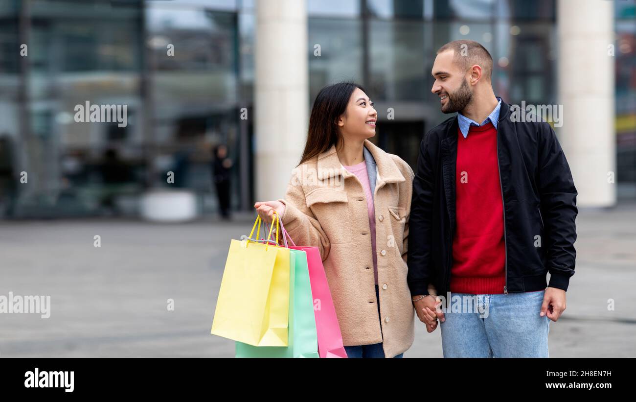 Fröhliches internationales Paar in warmen Outfits, das mit Einkaufstaschen auf der Straße in der Nähe eines großen Einkaufszentrums läuft, Kopierbereich Stockfoto