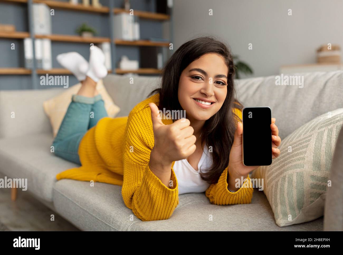 Sieh dir diese mobile App an. Glückliche arabische Dame demonstriert Smartphone mit leerem Bildschirm und zeigt Daumen nach oben Stockfoto
