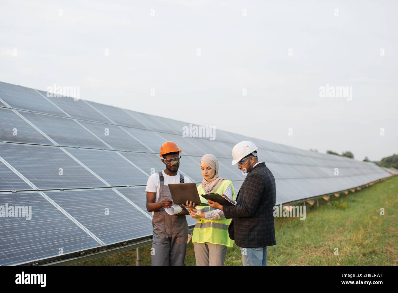 Muslimische Frau in Hijab und indischer Mann in weißem Helm im Gespräch mit afroamerikanischem Techniker auf Solarstation. Menschen mit mehreren Rassen, die Laptops, Clipboard und Blaupausen für die Arbeit im Freien verwenden. Stockfoto