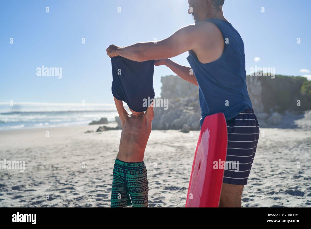 Vater hilft dem Sohn, das Hemd am sonnigen Sommerstrand zu entfernen Stockfoto