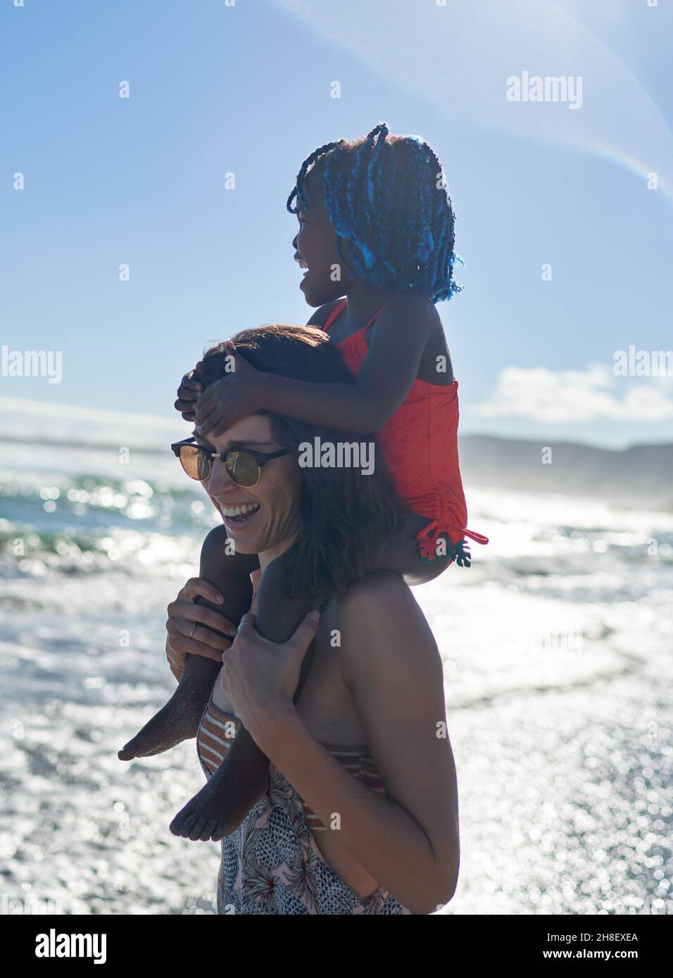 Glückliche Mutter trägt süße Tochter auf den Schultern am sonnigen Strand des Ozeans Stockfoto