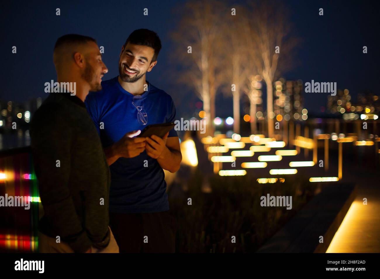 Glückliche Männer, die nachts im beleuchteten Stadtpark ein digitales Tablet nutzen Stockfoto