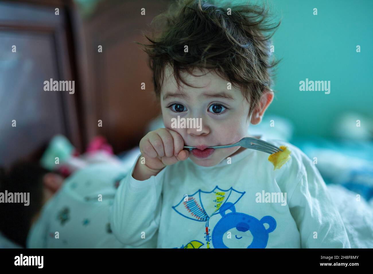 Kleiner Junge mit großen braunen Augen und unordentlichen braunen Haaren in weißem Schlafanzug mit einem blauen Bären, der einen Pfannkuchen mit einer Gabel frisst Stockfoto