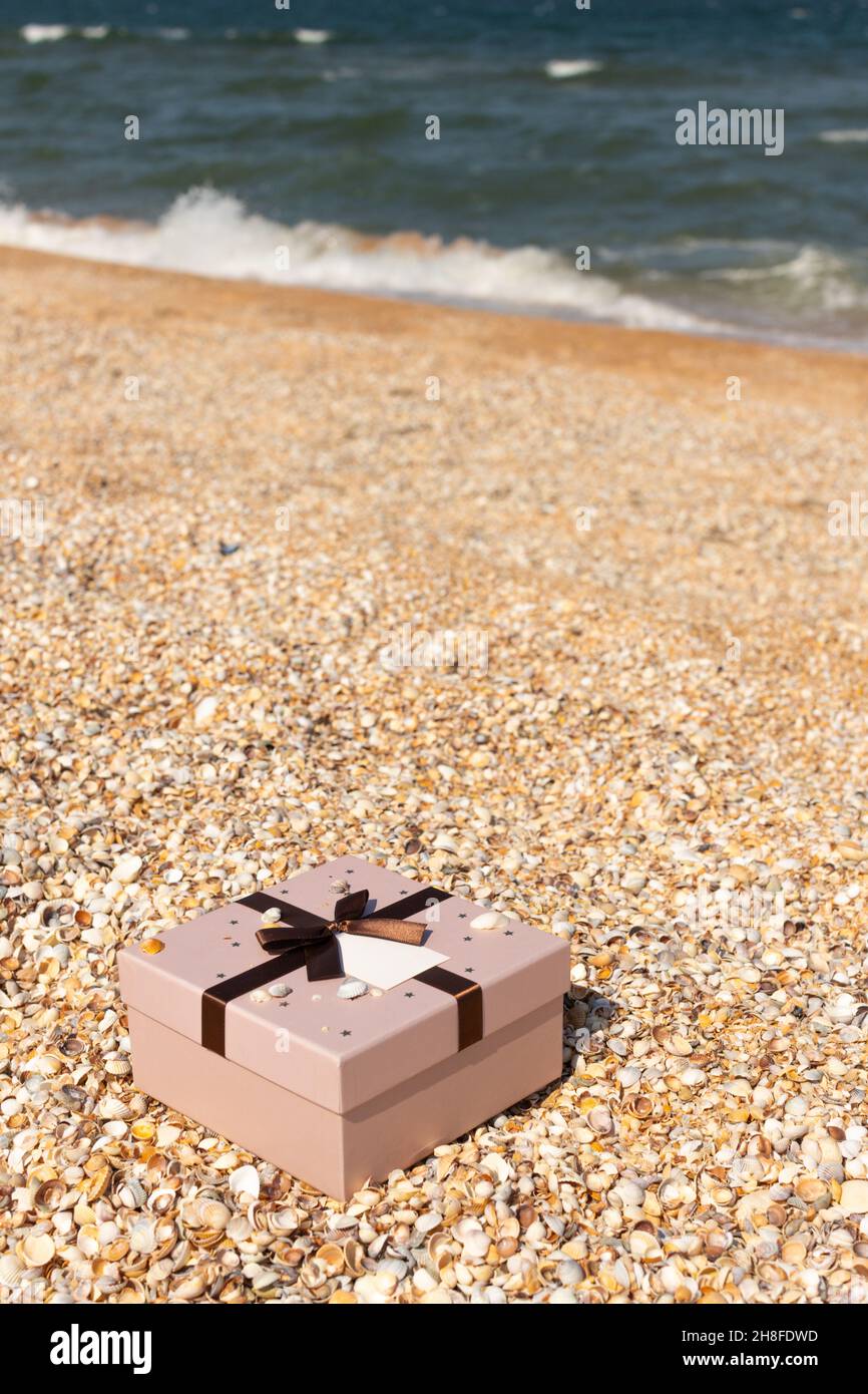 Am Strand in der Nähe des Meeres liegt eine Schachtel mit einem mit einem Band gebundenen Geschenk. Vorderansicht, vertikal. Stockfoto