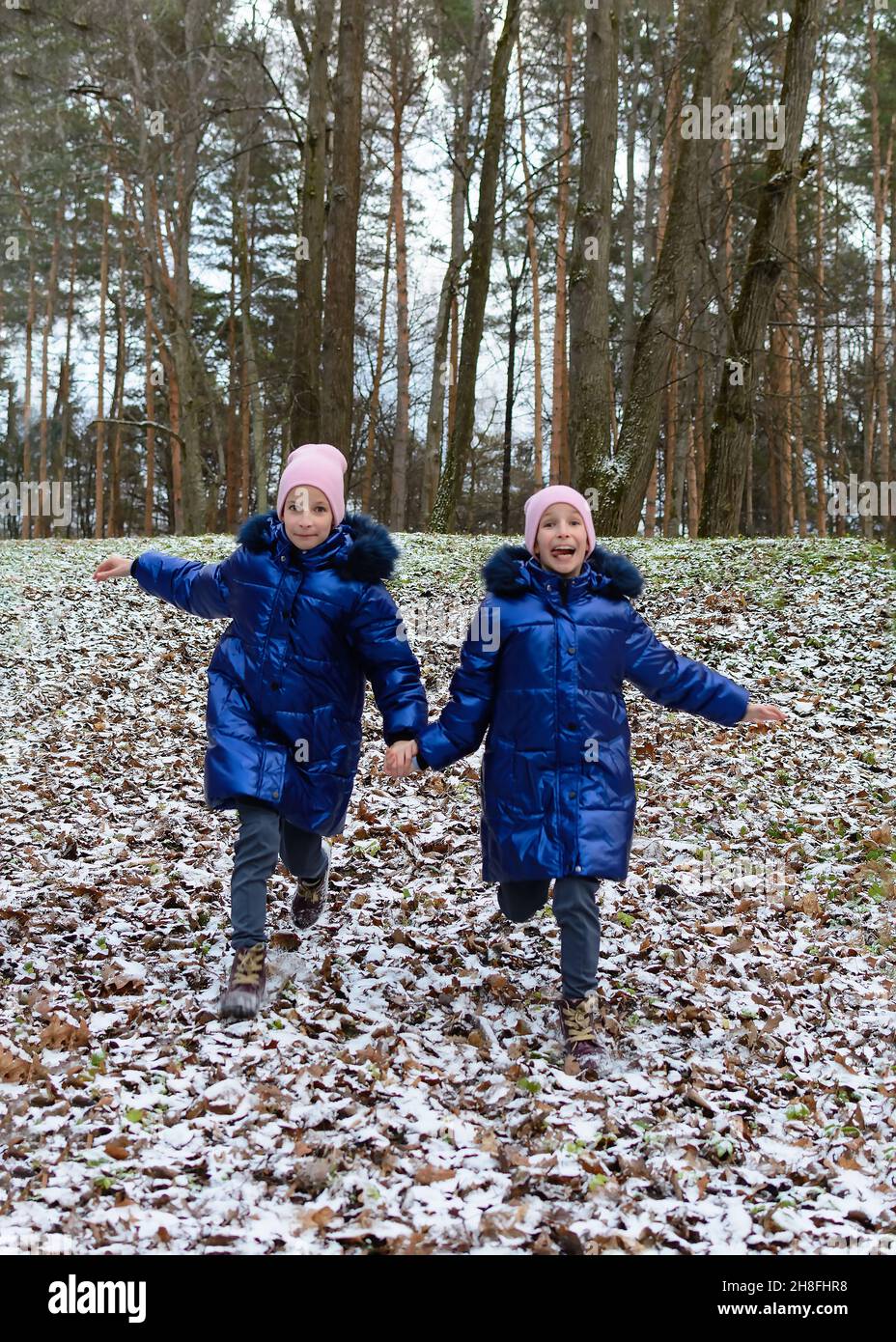 Der erste Frost im Herbst. Zwei glückliche Zwillingsmädchen in blauen Mänteln, Hand in Hand, die den Hügel hinunter laufen, bedeckt mit den trockenen Blättern und dem ersten Schnee im Vordergrund Stockfoto