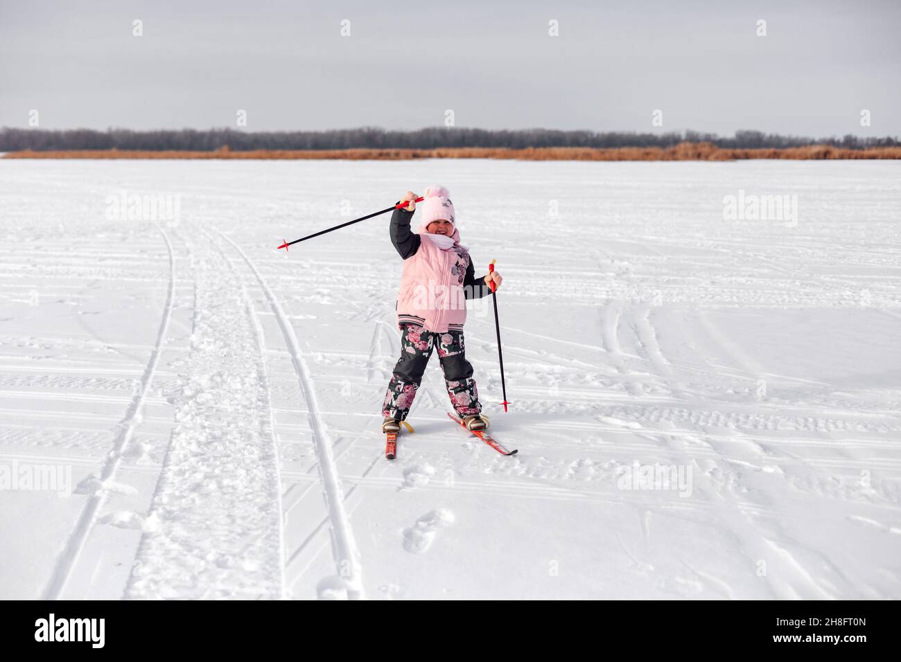 Kind fährt im Schnee Ski. Glückliches Mädchen im warmen Trainingsanzug lernt auf gefrorenem Flussufer Ski zu fahren und winkt ihre Hand, Winterlandschaft, verschneiten Hintergrund Stockfoto