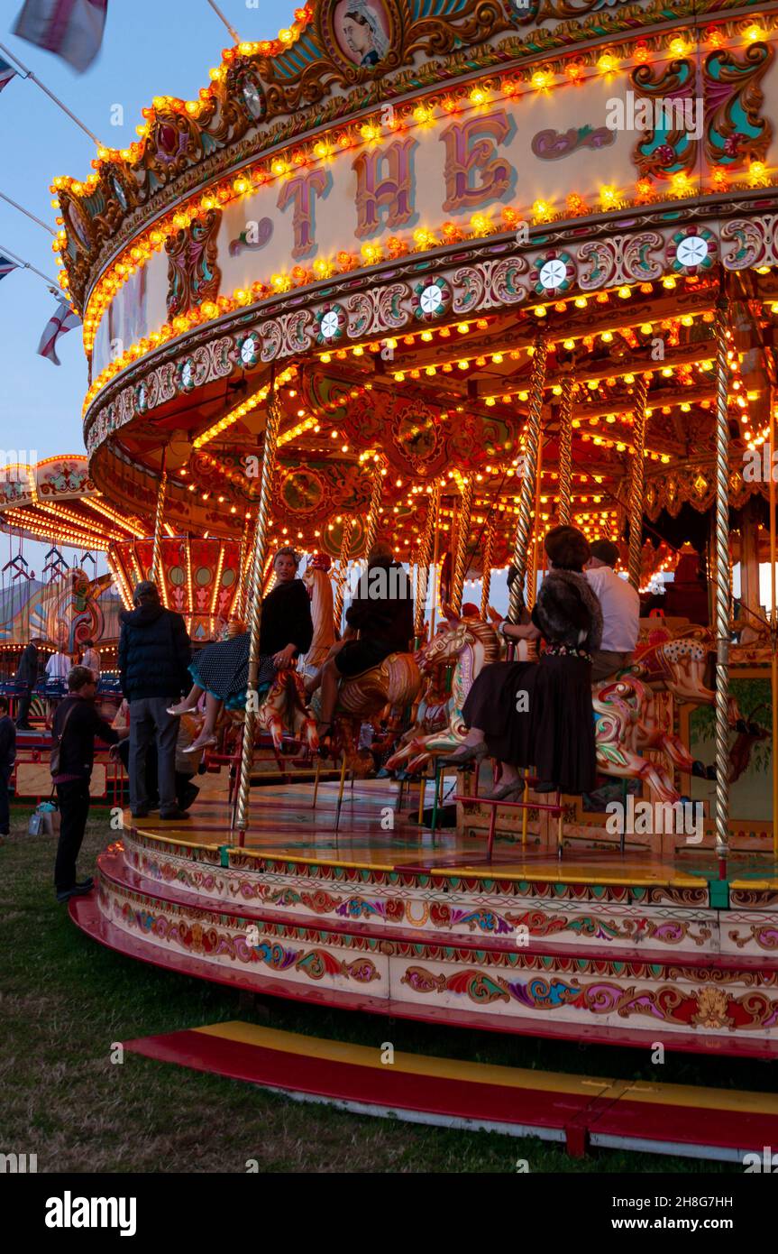 Classic Carousel, Merry Go Round, beim Goodwood Revival Vintage Event 2014, bei Dämmerung mit Lichtern. Karussell-Vergnügungsfahrt mit Menschen in Kostümen Stockfoto