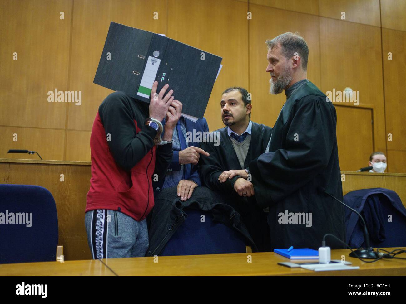 30. November 2021, Hessen, Frankfurt/Main: Mit einem Aktenordner vor seinem Gesicht, der Irakin Taha Al-J. (l) spricht am Morgen vor der Urteilsverkündung vor dem Oberlandesgericht Frankfurt mit seinen Anwälten Serkan Alkan (M) und Martin Heising (r). Die Staatsanwaltschaft wirft ihm Völkermord, Verbrechen gegen die Menschlichkeit, Kriegsverbrechen, Menschenhandel und Mord vor. Als mutmaßliches Mitglied der Terrormiliz IS soll er eine yezidische Frau und ihre Tochter als Sklaven festgehalten und missbraucht haben. Dabei soll er den fünfjährigen in Falludscha, Irak, gefesselt haben, wo das Kind an diesem starb Stockfoto