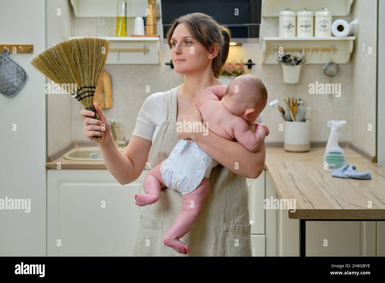 Eine Frau hält ein Kleinkind in ihren Armen, während sie in einer Küche zu Hause putzt, Probleme mit der Hausarbeit nach der Geburt eines Kindes Stockfoto