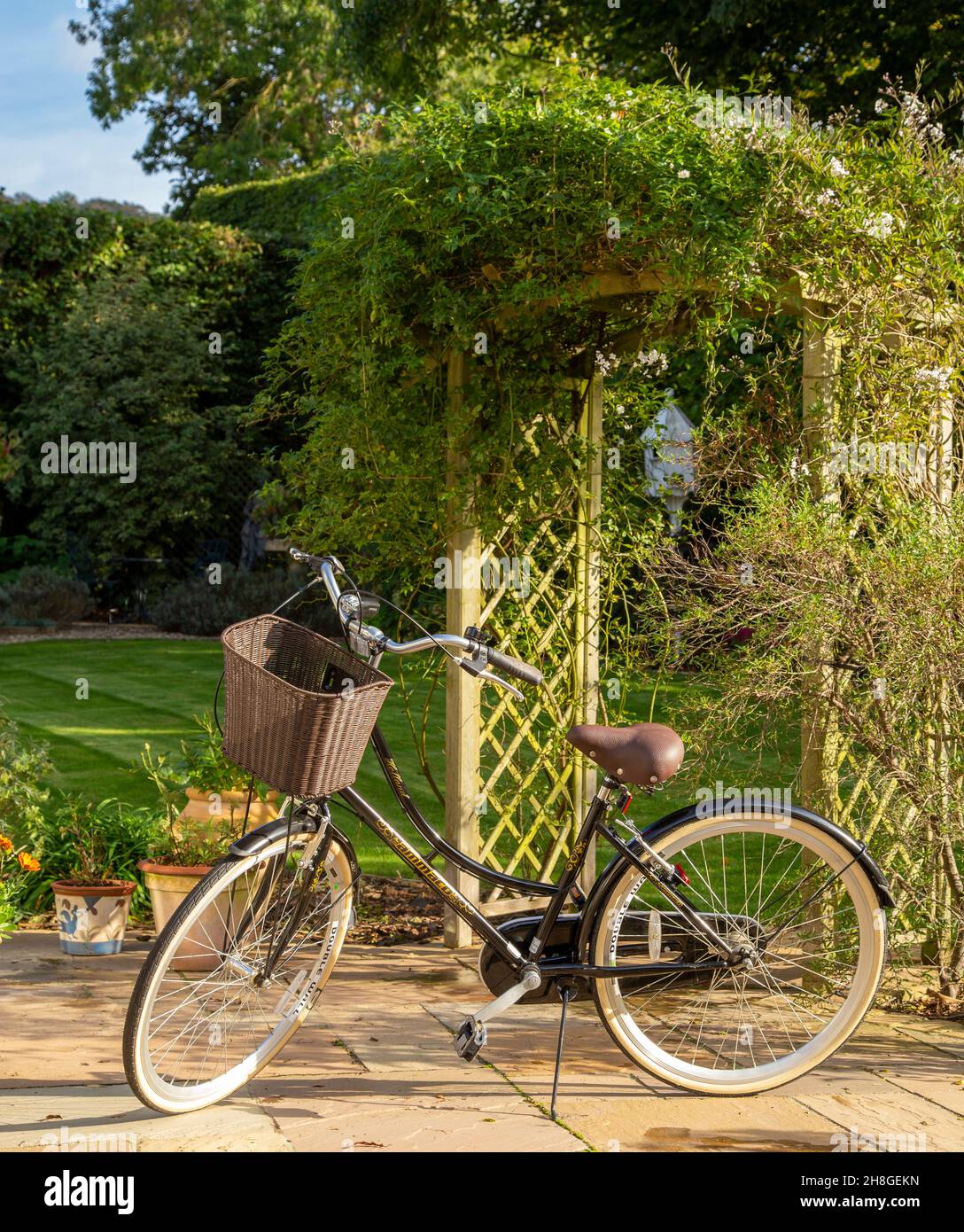 Das Ammaco 'Holland' Damenfahrrad im holländischen Stil befindet sich in einem hübschen Garten neben einem Bogen, mit Korb vor dem Lenker. Stockfoto