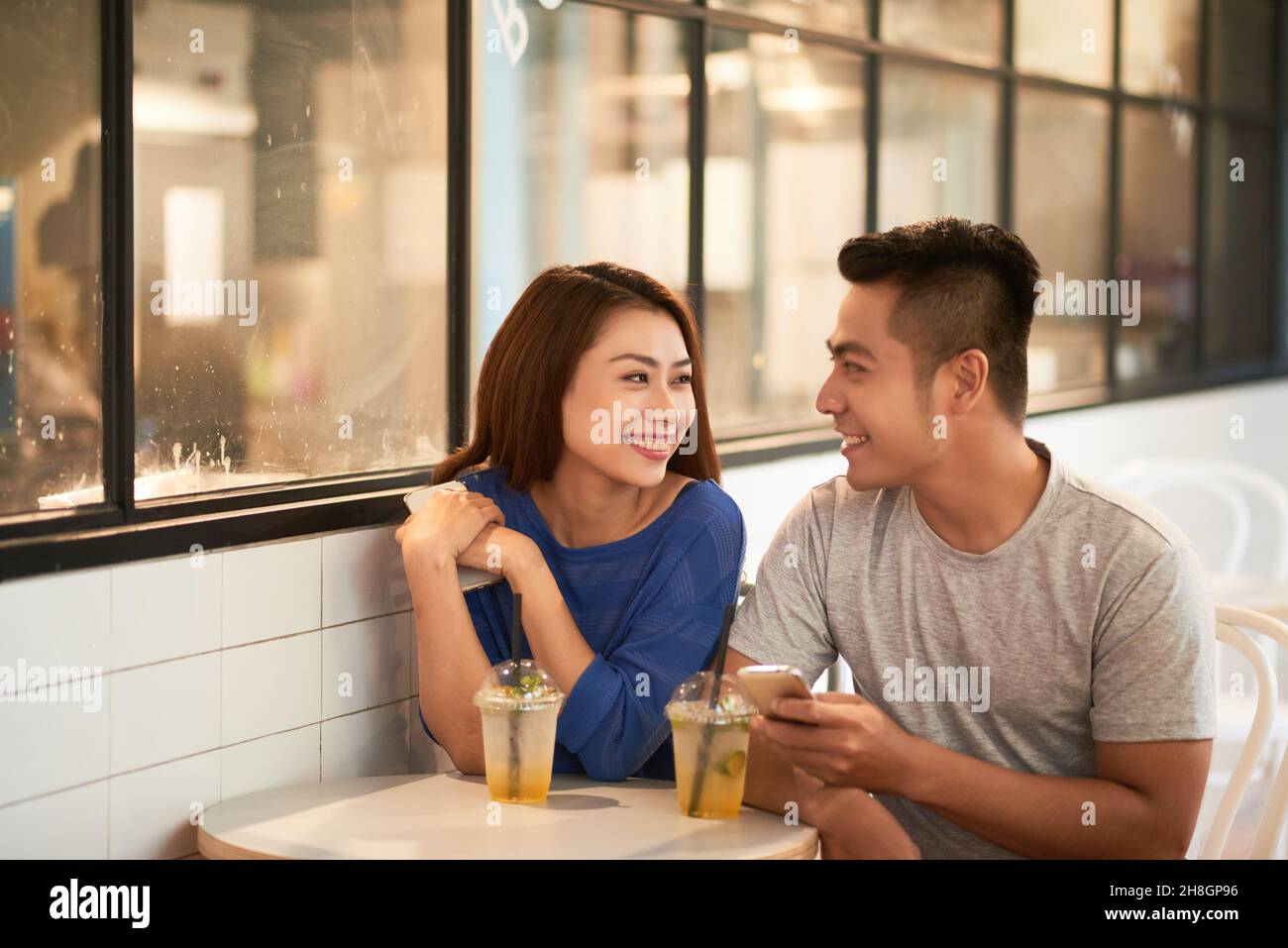 Lächelndes junges asiatisches Paar, das mit Smartphones an einem kleinen Tisch sitzt und im modernen Café spricht Stockfoto