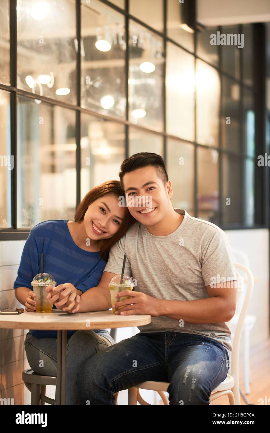Porträt eines glücklichen jungen asiatischen Paares, das mit Getränken an einem kleinen Tisch sitzt und die Hände hält Stockfoto