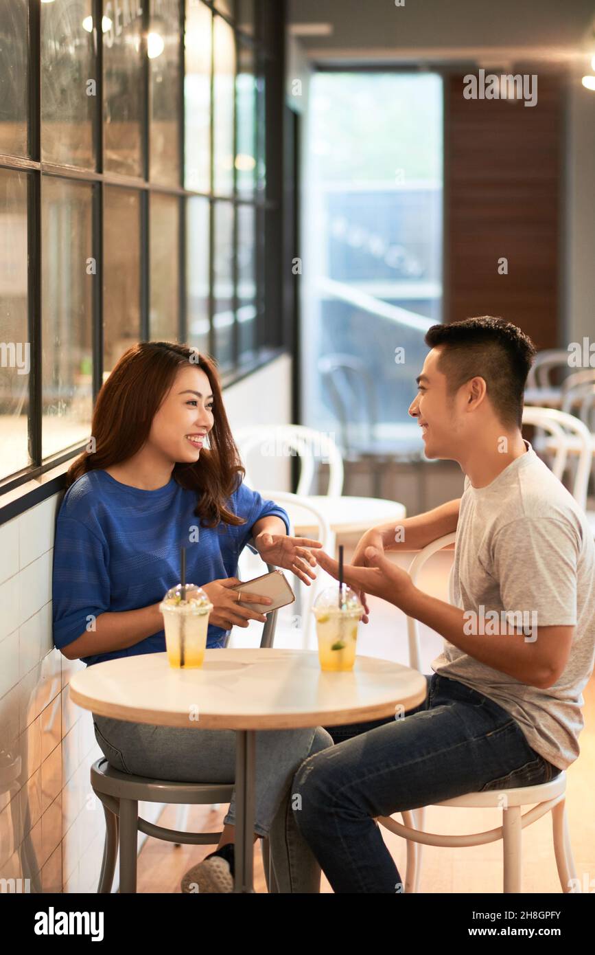 Lächelndes junges asiatisches Paar in lässigen Outfits, das bei einem Drink an einem kleinen Tisch sitzt und im Café miteinander plaudert Stockfoto