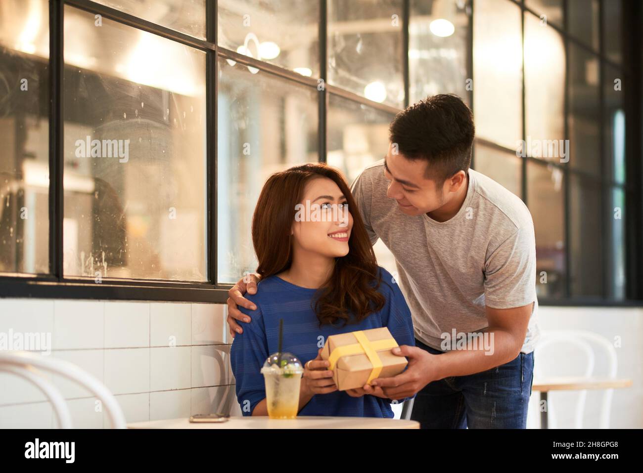 Liebevoller junger asiatischer Mann umarmt Freundin und gibt ihr Geschenk, während sie Überraschung im Café macht Stockfoto