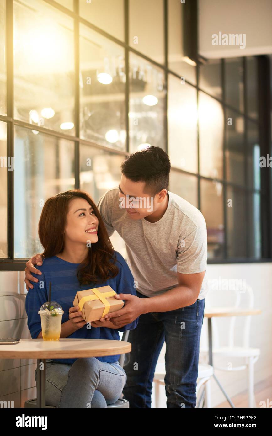 Glückliches junges asiatisches Paar in lässigen Outfits, das Jubiläum im Café feiert: Mann, der Freundin ein Geschenk gibt Stockfoto