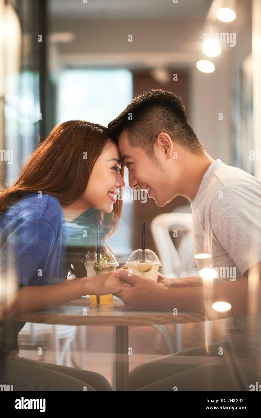 Lächelndes, anhängliches junges asiatisches Paar, das Hände hält und die Stirn berührt, während es im Café ein Date veranstaltet Stockfoto
