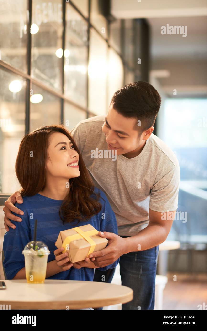 Lächelnder junger Mann umarmt asiatische Freundin von hinten im Café und gibt ihr ein Geschenk Stockfoto