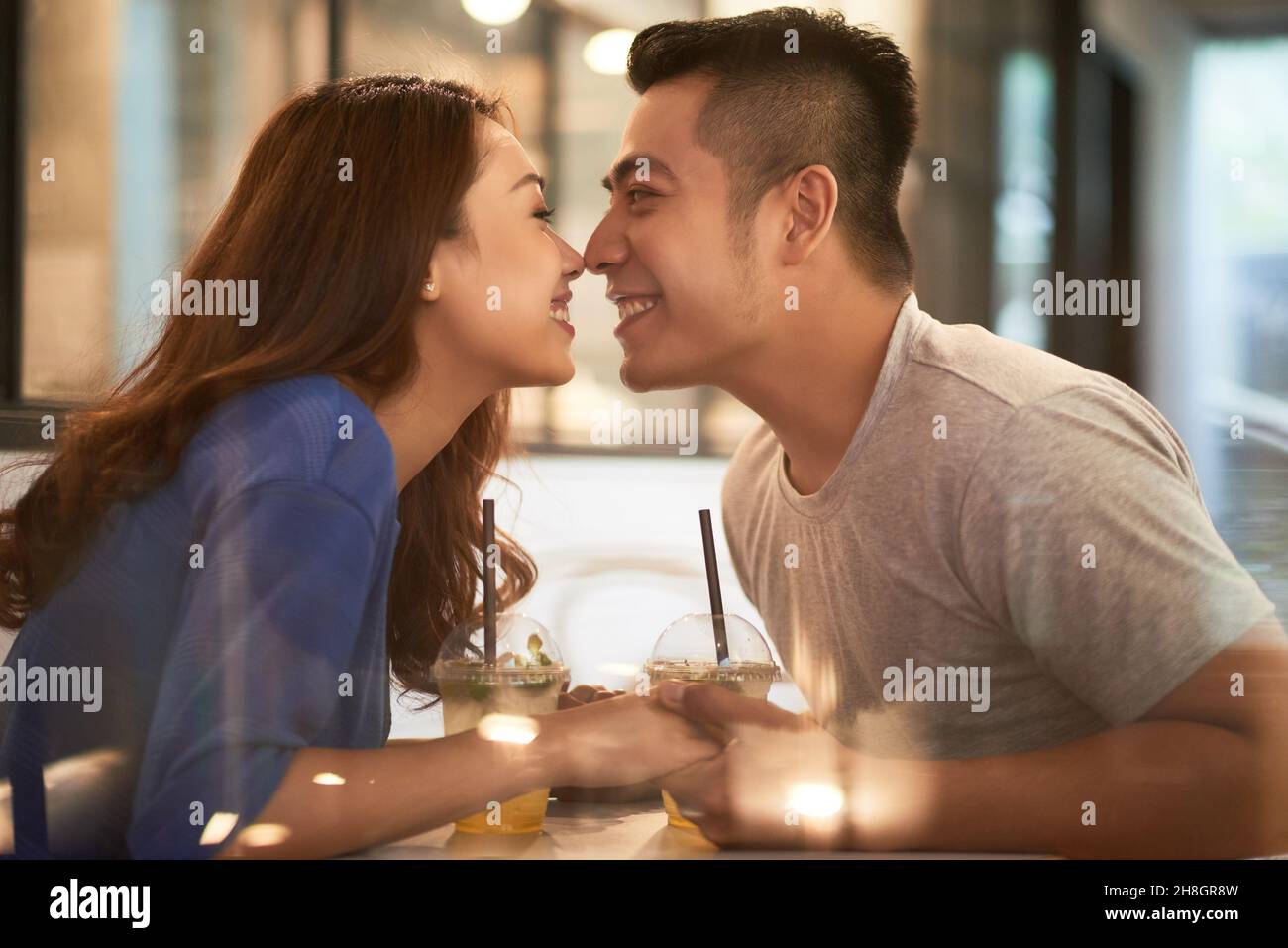 Fröhliches, liebevolles junges asiatisches Paar, das mit Sommercocktails und berührenden Nasen über dem Tisch im Café sitzt Stockfoto