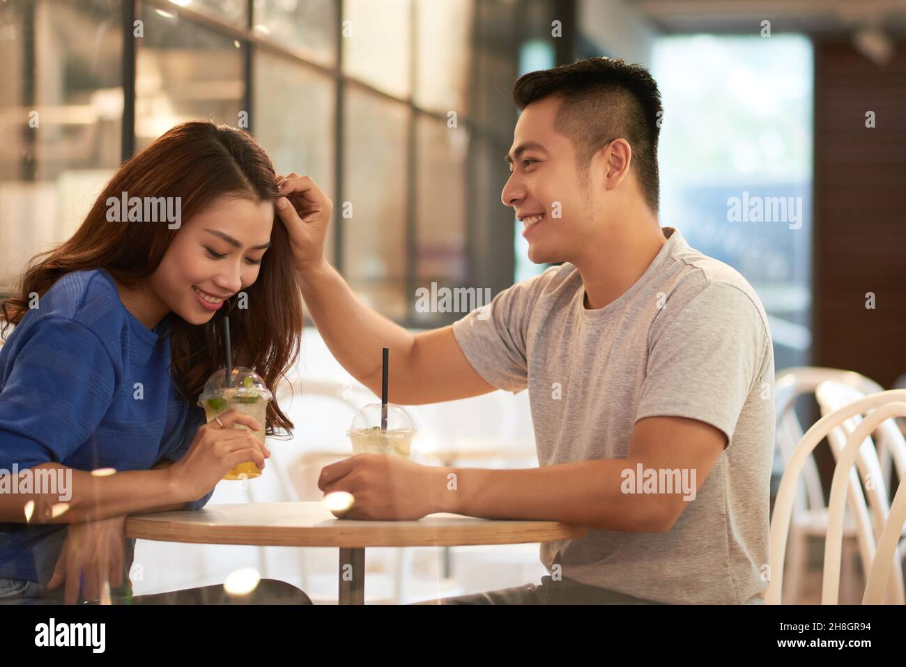 Lächelnder junger asiatischer Mann, der an einem kleinen runden Tisch sitzt und die Haare im Café an die Freundin anpasst Stockfoto