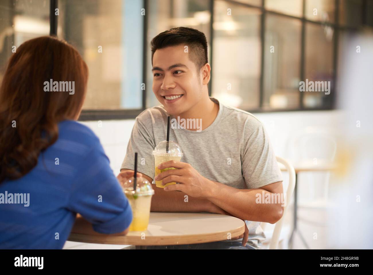 Lächelnder junger asiatischer Mann im T-Shirt, der am Tisch sitzt und Limonade trinkt, während er sich mit dem Mädchen im Café vertrunken hat Stockfoto