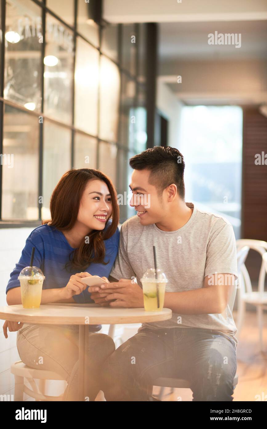 Lächelndes, schönes junges asiatisches Paar, das an einem kleinen runden Tisch mit erfrischenden Cocktails im Café sitzt und durch das Smartphone scrollt Stockfoto