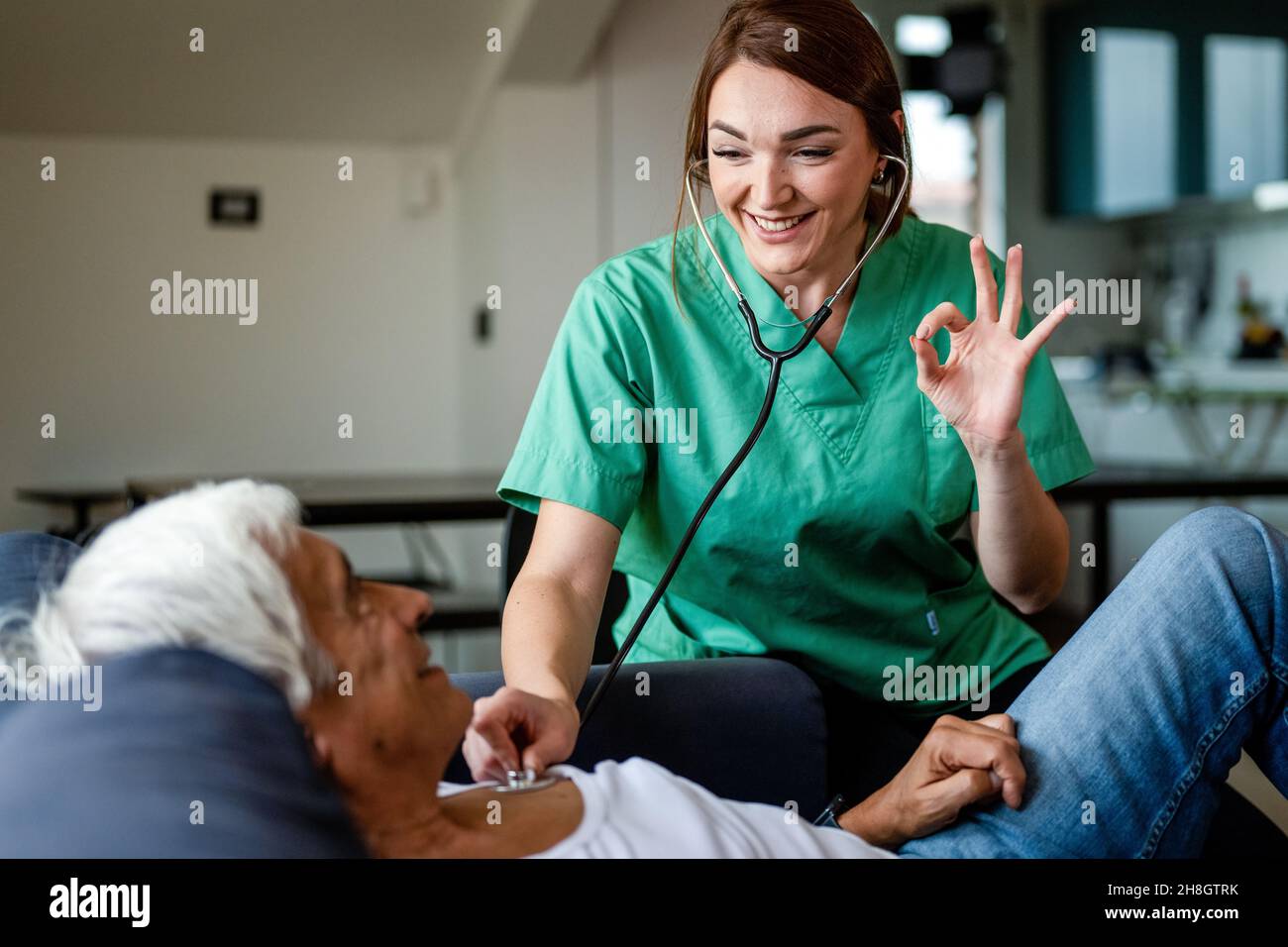 Gesundheitsbesucher, der Stethoskop verwendet, um die Gesundheit eines älteren Mannes zu überprüfen, junge Krankenschwester, die während eines Hausbesuchs mit der Hand in Ordnung gestikuliert, Konzept der häuslichen Pflege Stockfoto