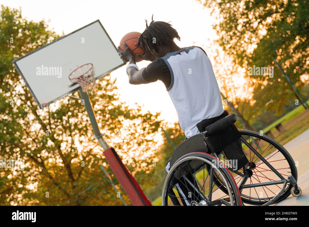 Junger Athlet auf Rollstuhl wirft Ball in Basketball-Korb, Sonnenuntergang-Effekt im Hintergrund, soziale incluzion und sortieren Vielfalt Konzept Stockfoto