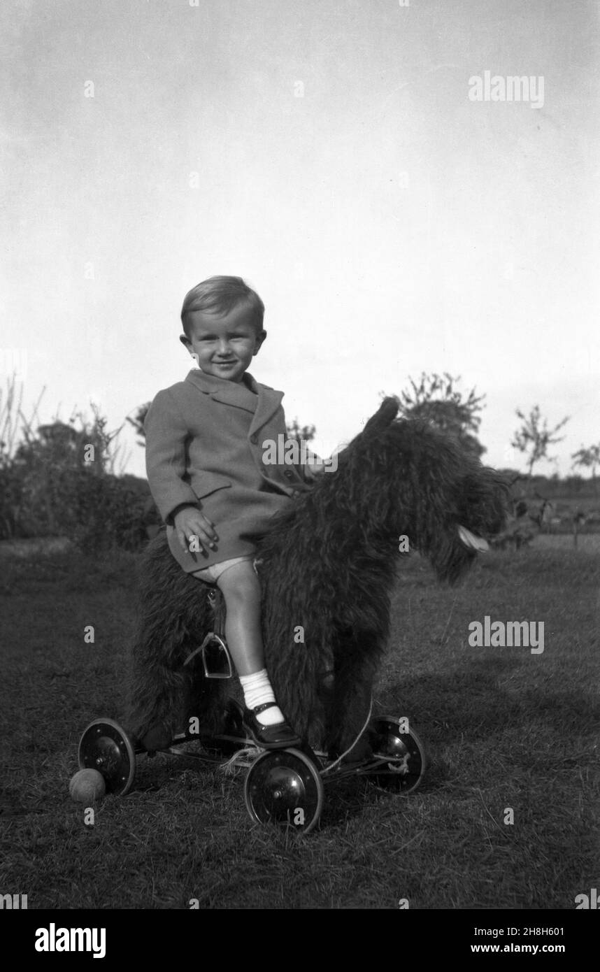 1950s, historisch, draußen auf etwas Gras, ein kleiner Junge, der für ein Foto auf einem großen pelzigen Spielzeughund auf Rädern sitzt, England, Großbritannien. Ride-On oder Push-Along-Spielzeuge mit Rädern waren in dieser Zeit ein beliebtes Kinderspielzeug Stockfoto