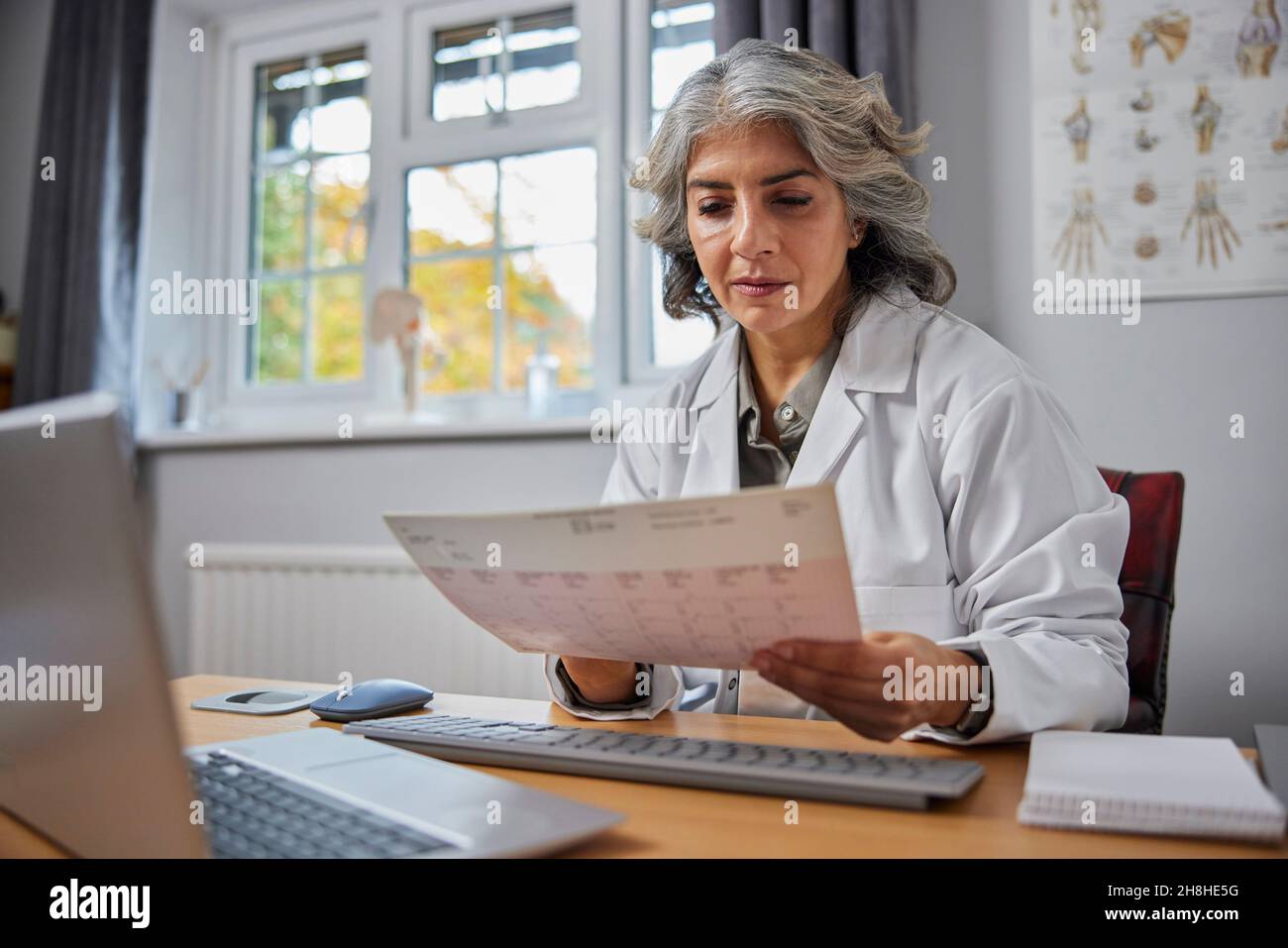 Ältere Frau GP trägt weißen Mantel am Schreibtisch in Arztpraxis Blick auf EKG-Anzeige Stockfoto