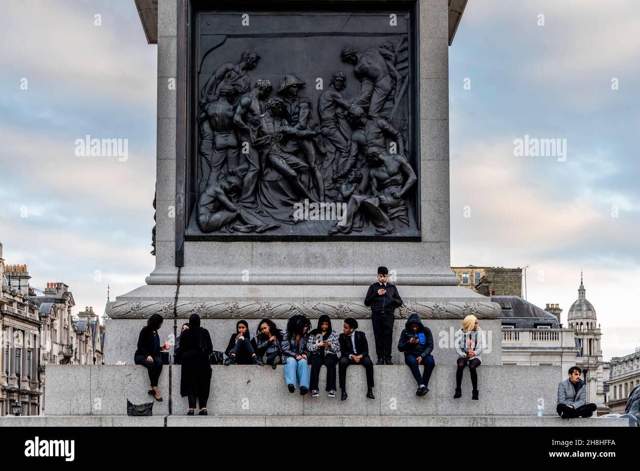 Eine multiethnische Gruppe junger Menschen, die auf den Stufen der Nelson-Säule, Trafalgar Square, London, Großbritannien, sitzt. Stockfoto