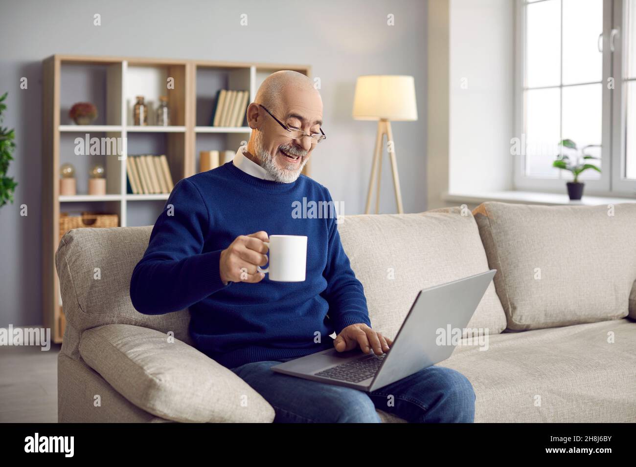 Glücklicher älterer Mann, der auf der Couch saß, seinen Laptop benutzte, Kaffee trank und lachte Stockfoto