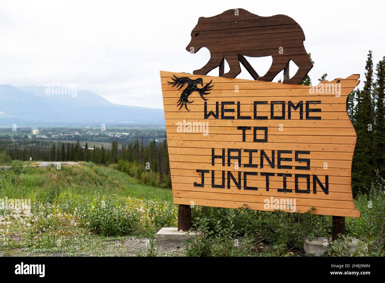 Schild für Haines Junction im Südwesten des Yukon, Kanada. Das Holzschild zeigt eine Figur, die einen Grizzlybären darstellt. Stockfoto