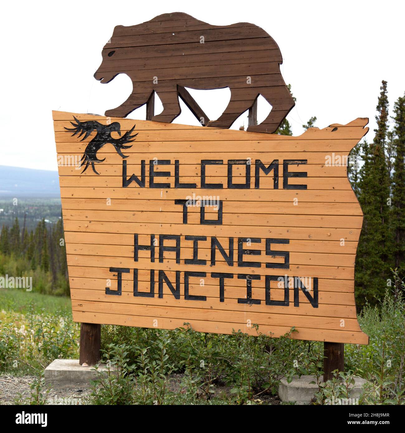 Schild für Haines Junction im Südwesten des Yukon, Kanada. Das Holzschild zeigt eine Figur, die einen Grizzlybären darstellt. Stockfoto