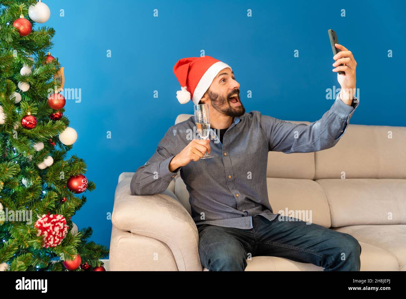 Der junge Mann feiert Weihnachten allein zu Hause, sitzt auf dem Sofa mit einem Getränk und lächelt am Telefon Stockfoto