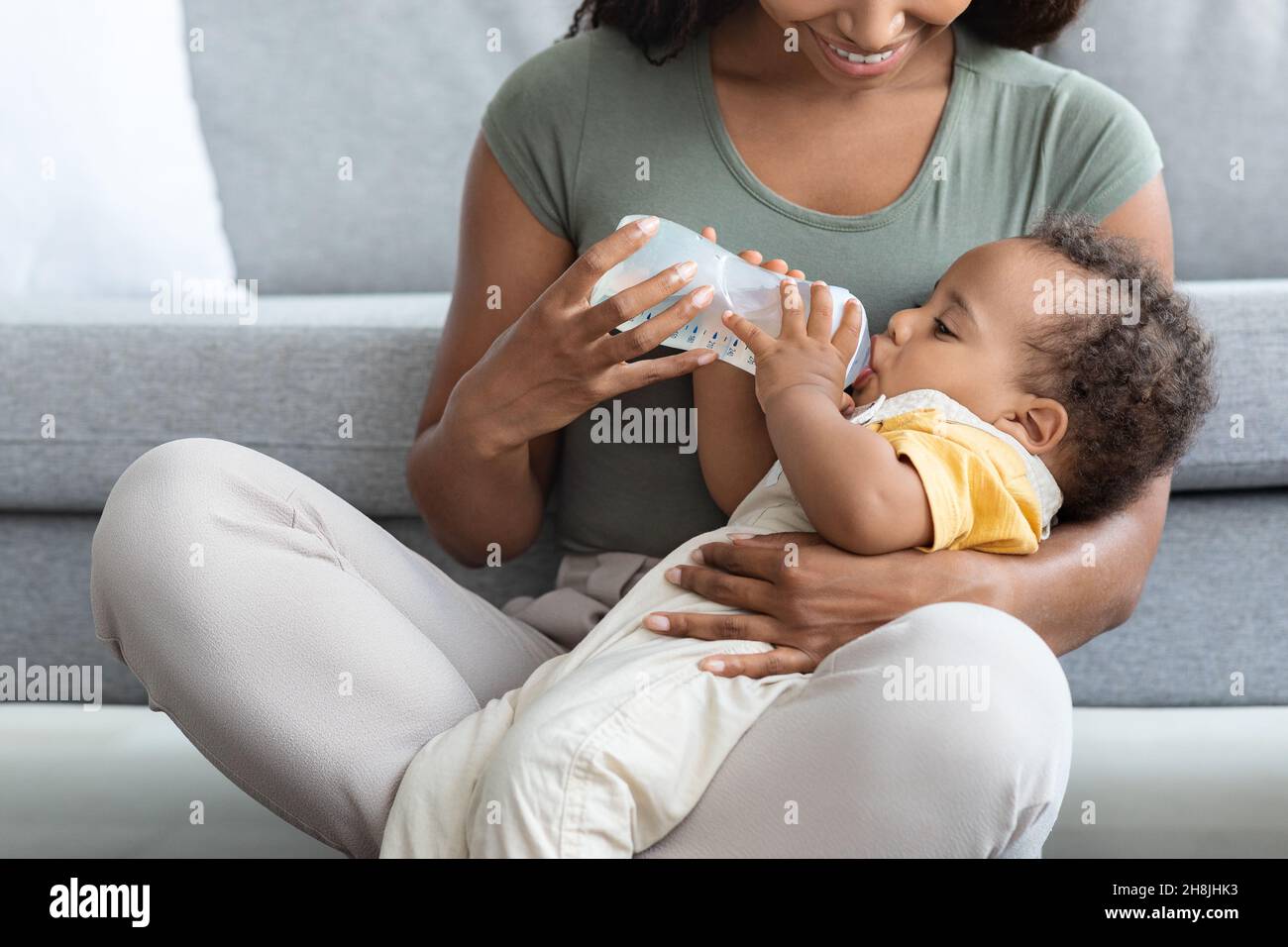 Schwarzes Baby trinkt Wasser aus der Flasche, während es sich auf Mutters Schoß entspannt Stockfoto