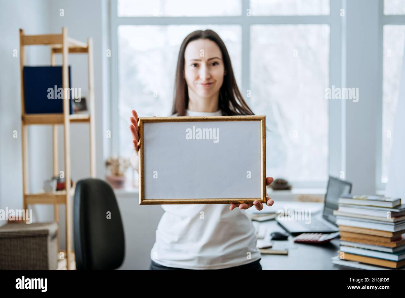 Rahmenbild, Plakat, Diplom, Urkunde in weiblichen Händen. Junge Brünette Frau hält leeren leeren hölzernen Fotorahmen auf Tisch mit Laptop Stockfoto